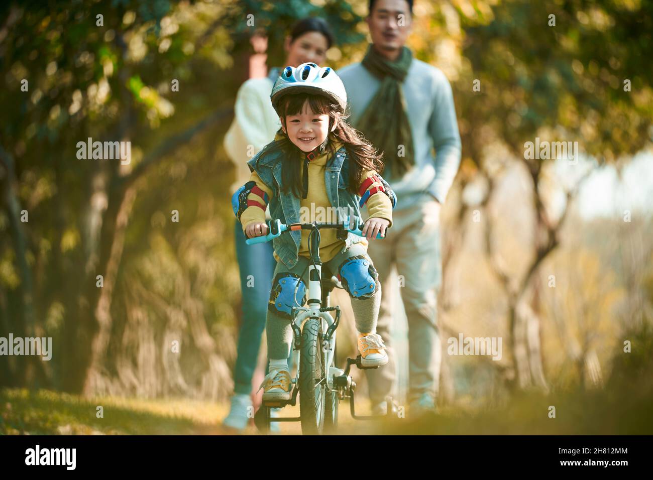 petite fille asiatique avec casque et équipement de protection complet à vélo dans le parc de la ville avec les parents regardant de l'arrière Banque D'Images