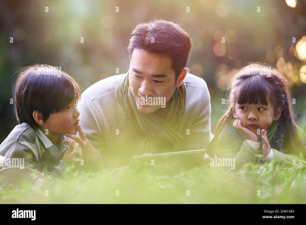 père asiatique couché sur le front de l'herbe racontant l'histoire à deux enfants heureux et souriants Banque D'Images