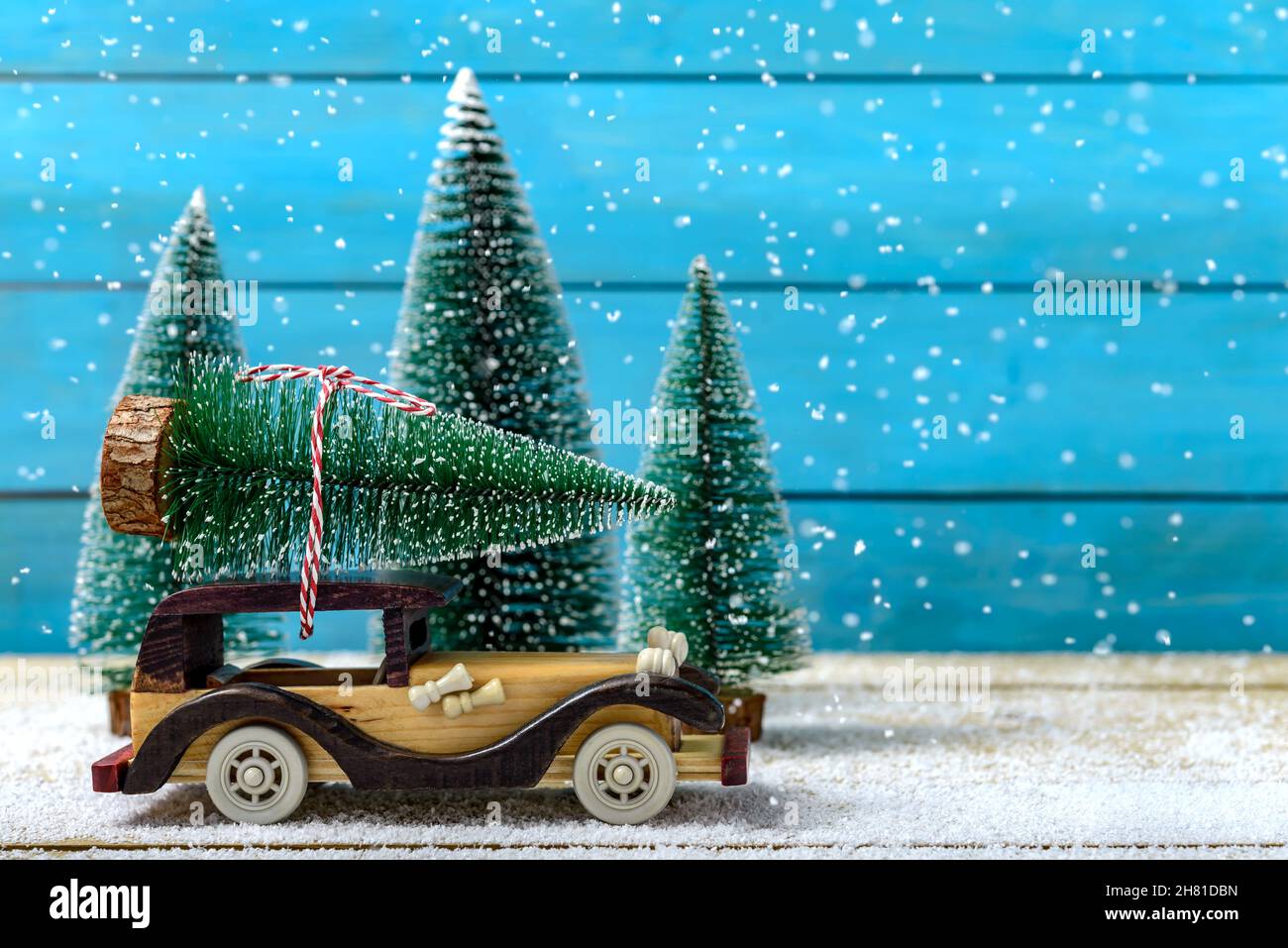 Voiture jouet avec arbre de Noël sur le toit à côté des sapins pendant qu'il neige sur une table en bois.Concept de célébration de Noël et du nouvel an. Banque D'Images