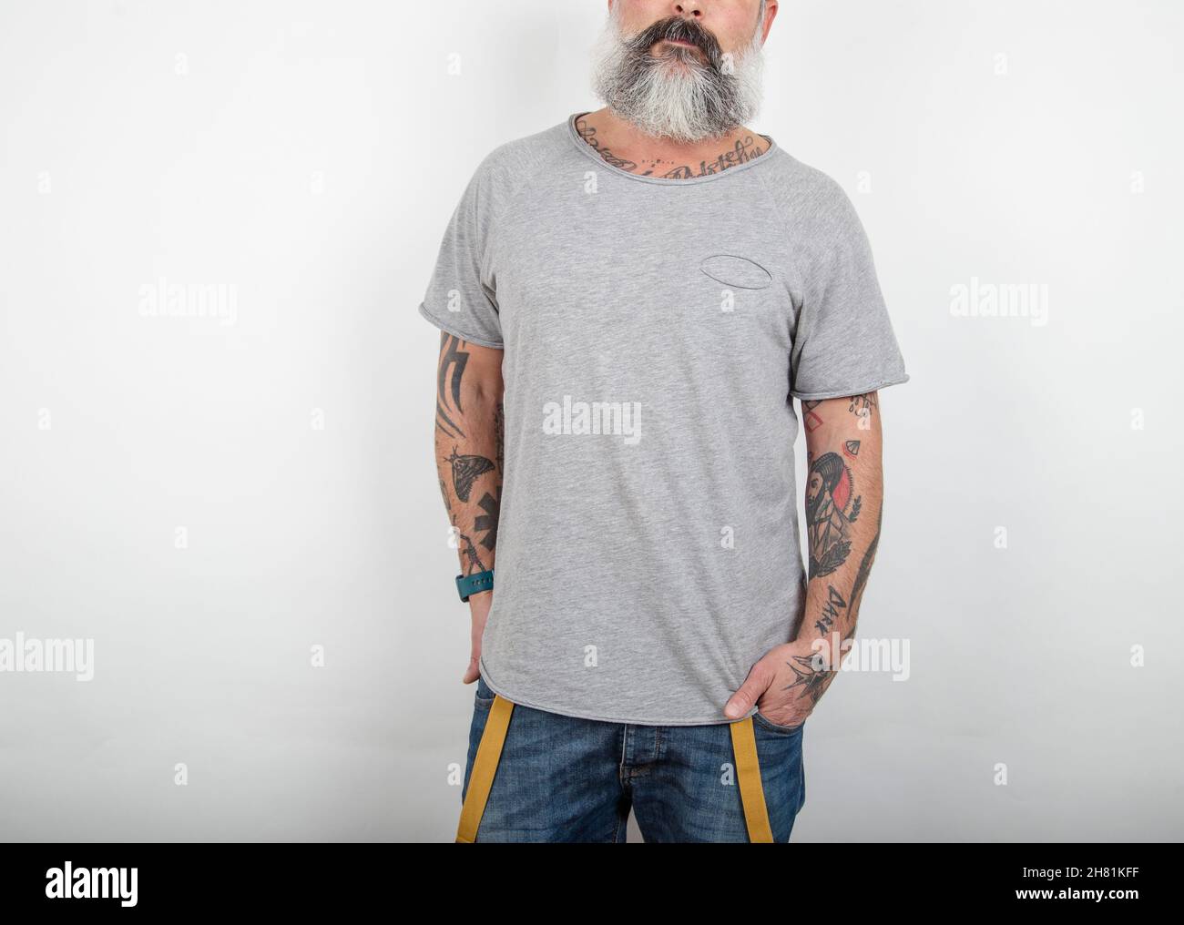 Gros plan d'un homme tatoué à barbe pose dans un Jean avec bretelles et un t-shirt en coton gris sur fond blanc. Banque D'Images