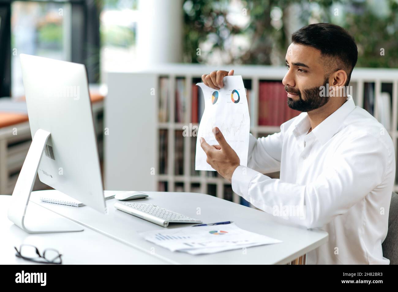 Homme d'affaires ou PDG indien sérieux et intelligent, avec une barbe au bureau, un ordinateur, un dialogue vidéo avec des partenaires d'affaires ou des employés, des documents et des graphiques Banque D'Images