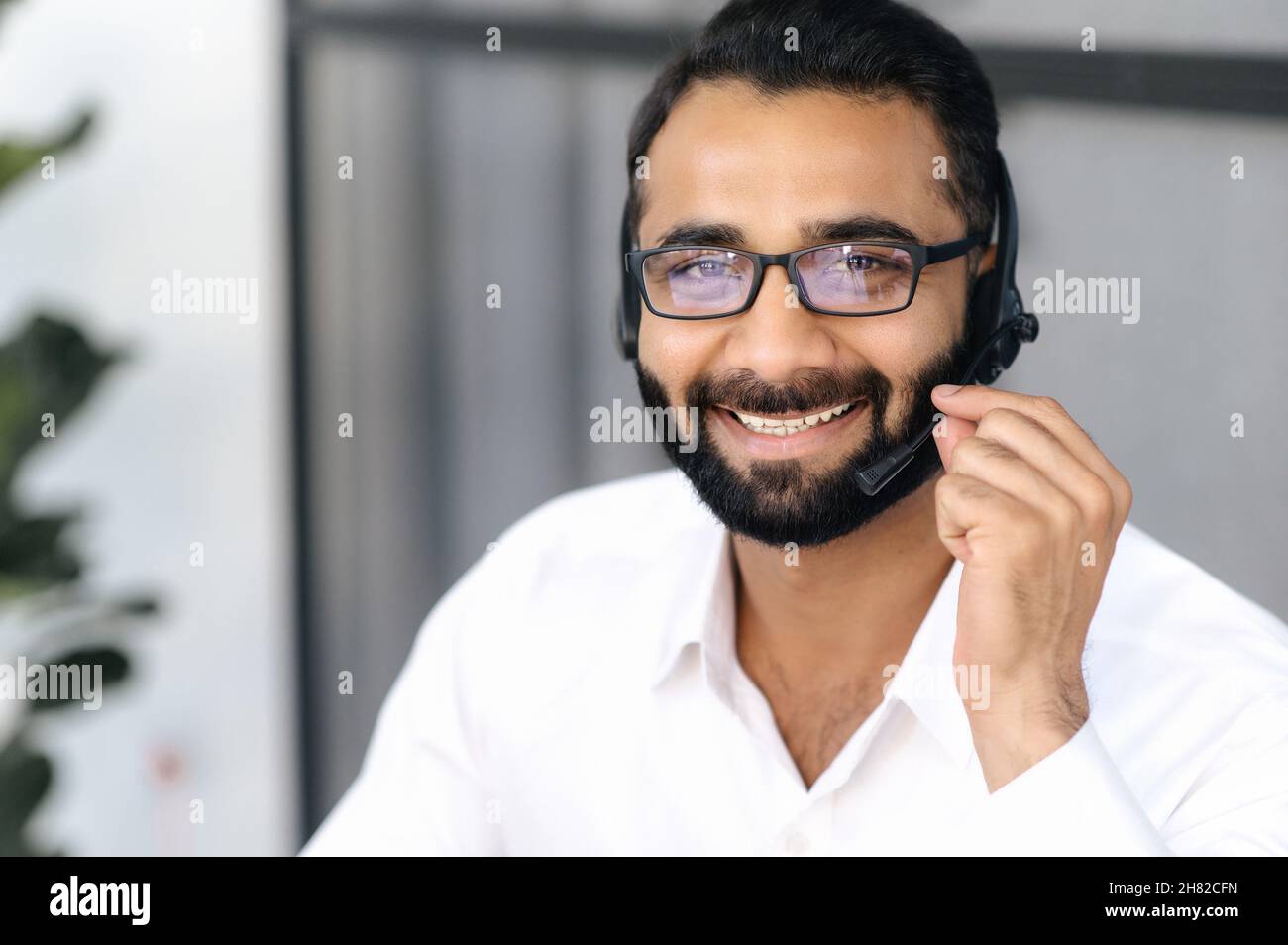 Gros plan d'un homme d'affaires indien intelligent, d'un opérateur de centre d'appels ou d'un consultant, portant des lunettes, utilisant un casque pour les conférences téléphoniques, regardant l'appareil photo et souriant amical Banque D'Images