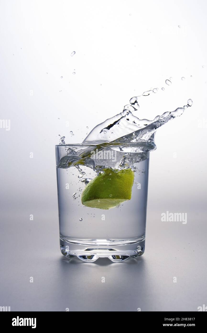 Morceau de chaux éclaboussant dans un verre de liquide clair ou d'eau sur une surface grise et un fond blanc Banque D'Images