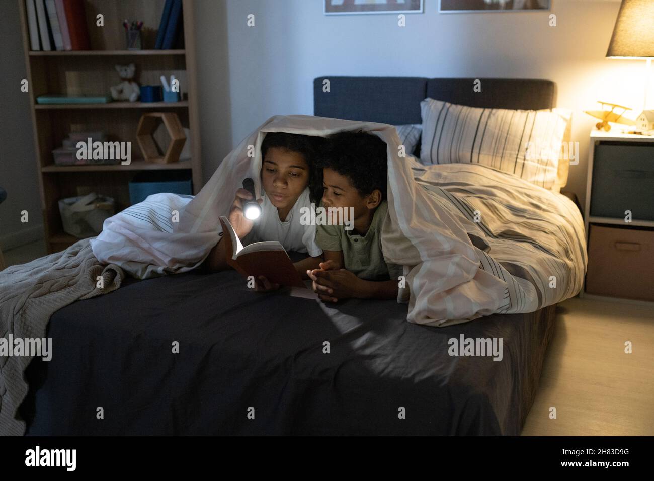 Soeur afro-américaine tenant une lampe de poche et un livre de lecture à frère pendant qu'ils sont au lit la nuit Banque D'Images