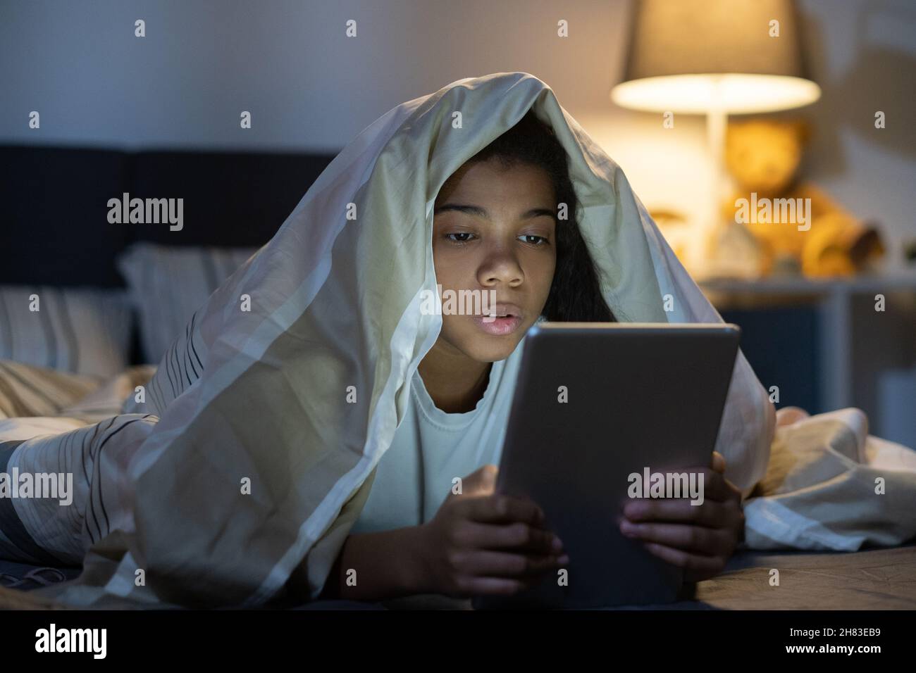 Curieuse fille afro-américaine couchée sous couette dans le lit et filet de surf sur tablette la nuit Banque D'Images