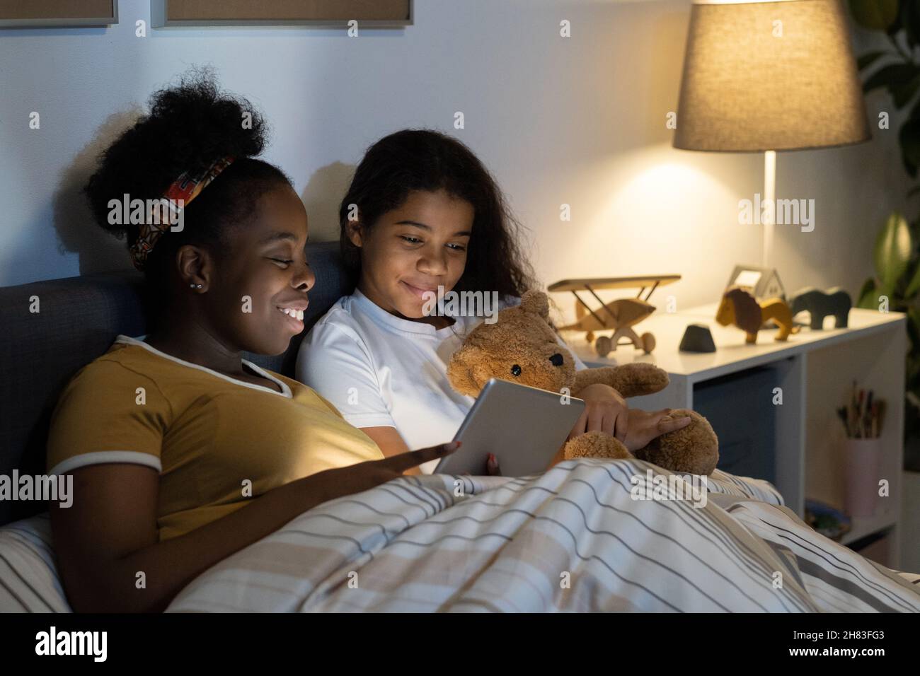 Jeune mère afro-américaine souriante en foulard regardant un film sur tablette avec une fille adolescente la nuit Banque D'Images