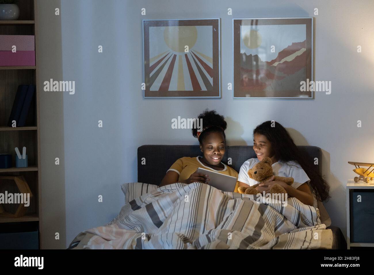 Une mère afro-américaine positive assise avec sa fille au lit et regardant un film sur une tablette dans une pièce sombre Banque D'Images