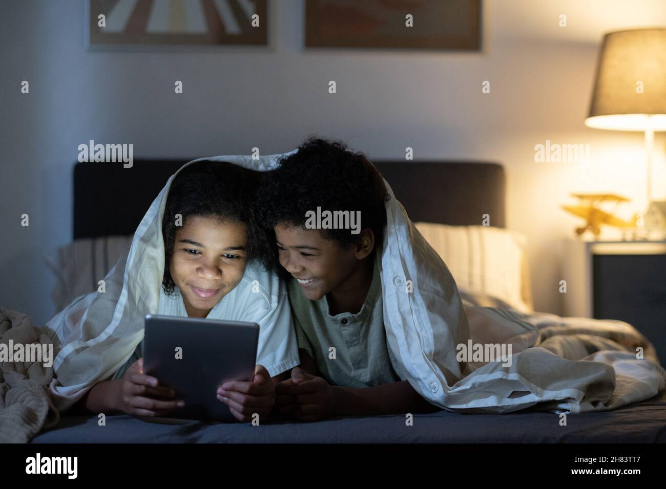 Frères et sœurs noirs positifs, couchés sous une couette sur le lit et utilisant une tablette tout en naviguant sur Internet la nuit Banque D'Images