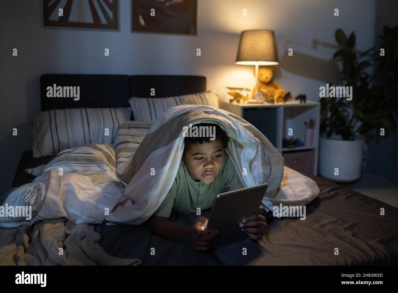 Garçon noir concentré couché sous couette et utilisant une tablette dans une chambre de bébé sombre Banque D'Images