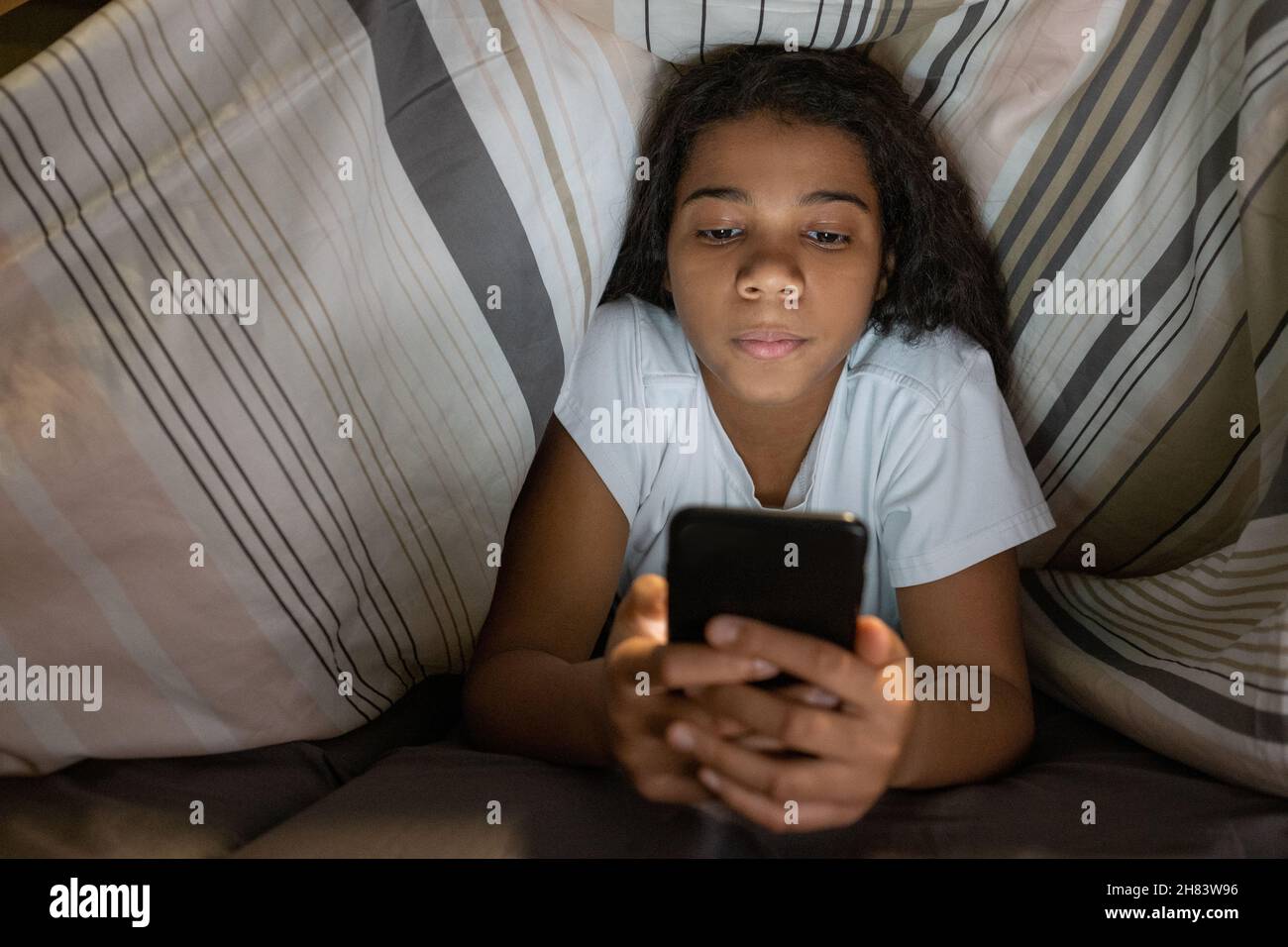 Contenu afro-américaine adolescente couchée sous une couette dans le lit et défilant à travers le smartphone Banque D'Images