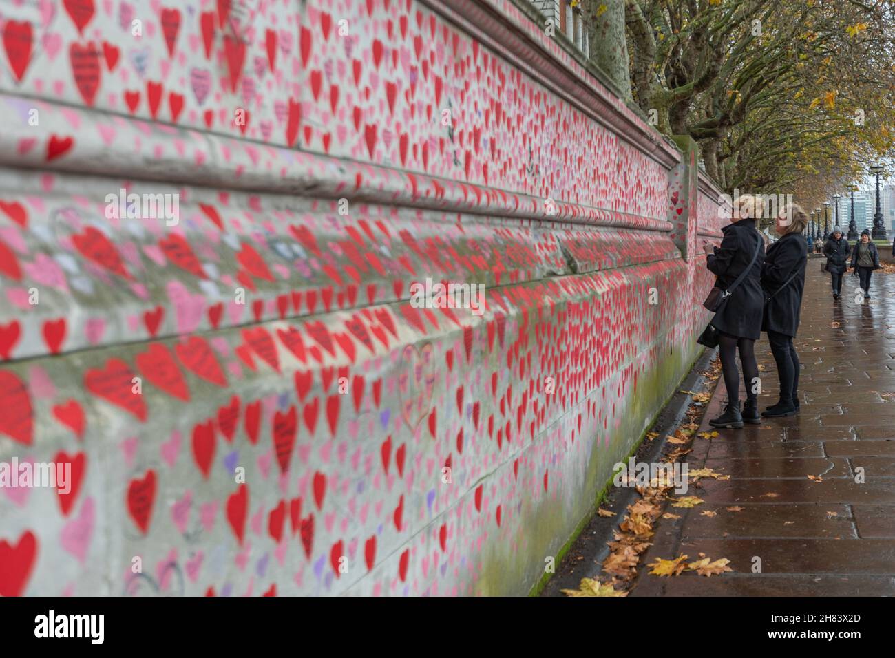 Londres, Royaume-Uni. 27 novembre 2021. Un long tronçon du National COVID Memorial Wall sur la rive sud de la Tamise, à Londres, recouvert de milliers de cœurs rouges et roses peints à la main. Chaque cœur représente une vie perdue par COVID-19 au Royaume-Uni. Deux individus se tiennent dans une réflexion tranquille. La chaussée humide et les feuilles d'automne suggèrent un jour pluvieux de souvenir. Penelope Barritt/Alamy Live News Banque D'Images