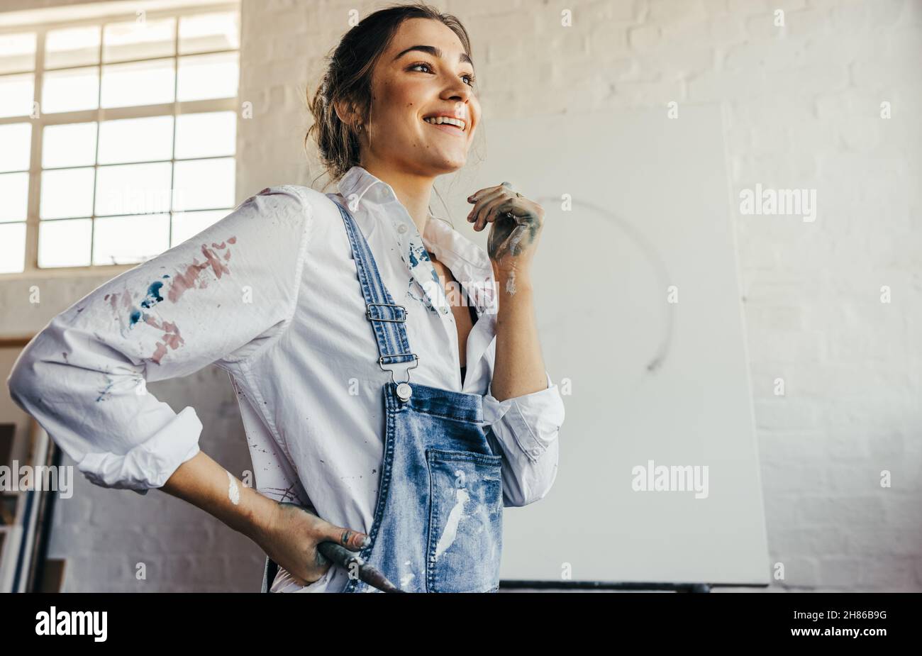 Femme peintre attentiste souriante dans son studio.Un jeune artiste contemplant de nouvelles idées créatives tout en se tenant devant une toile vierge.Imaginatif Banque D'Images