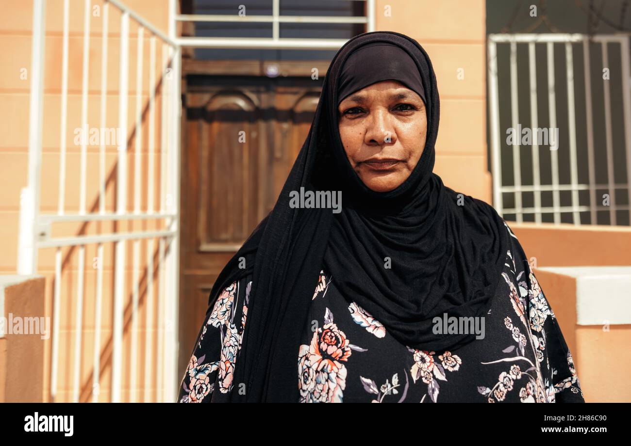 Femme musulmane confiante regardant la caméra tout en se tenant devant sa maison pendant la journée.Une femme d'âge moyen confiante porte un hijab et Banque D'Images Femme musulmane confiante regardant la caméra tout en se tenant devant sa maison pendant la journée.Une femme d'âge moyen confiante porte un hijab et Banque D'Images