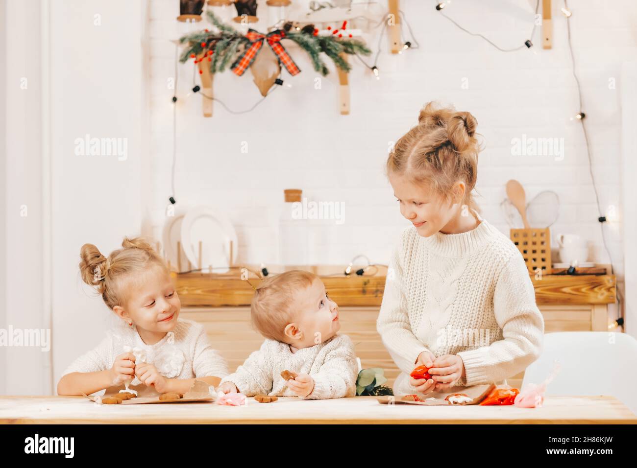 3 filles blondes décorent les biscuits de Noël avec du glaçage sur une table en bois dans la cuisine.Les amies cuisent ensemble dans la cuisine avec le Christ Banque D'Images