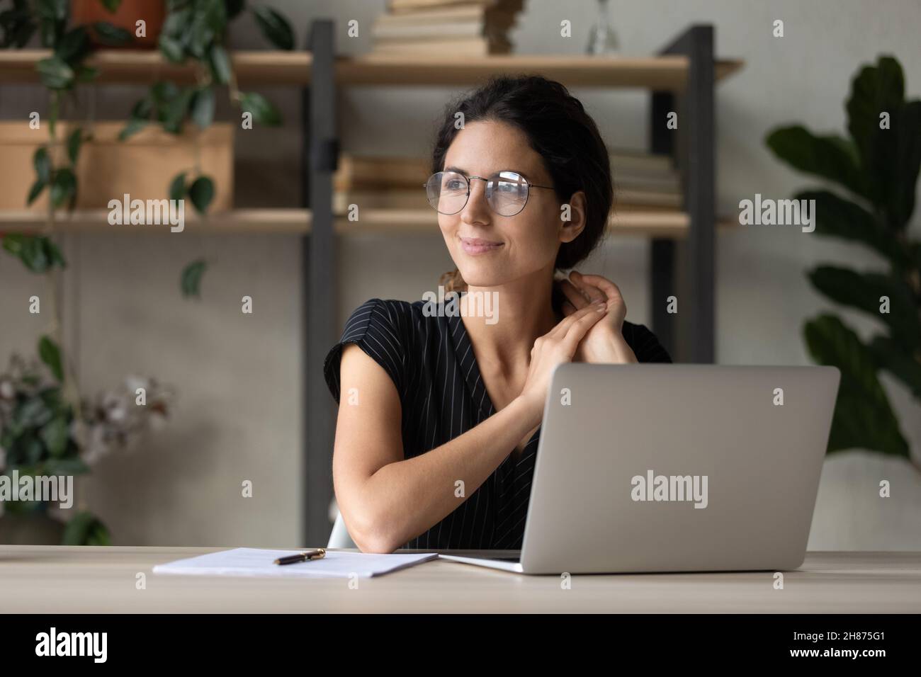 Une femme en latina souriante s'assoit sur un lieu de travail distrait de l'écran d'un ordinateur portable Banque D'Images
