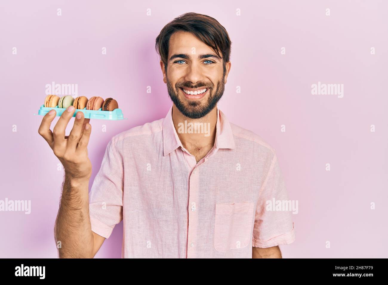Jeune homme hispanique portant de délicieuses pâtisseries de macrons, qui a l'air positif et heureux debout et souriant avec un sourire confiant montrant des dents Banque D'Images