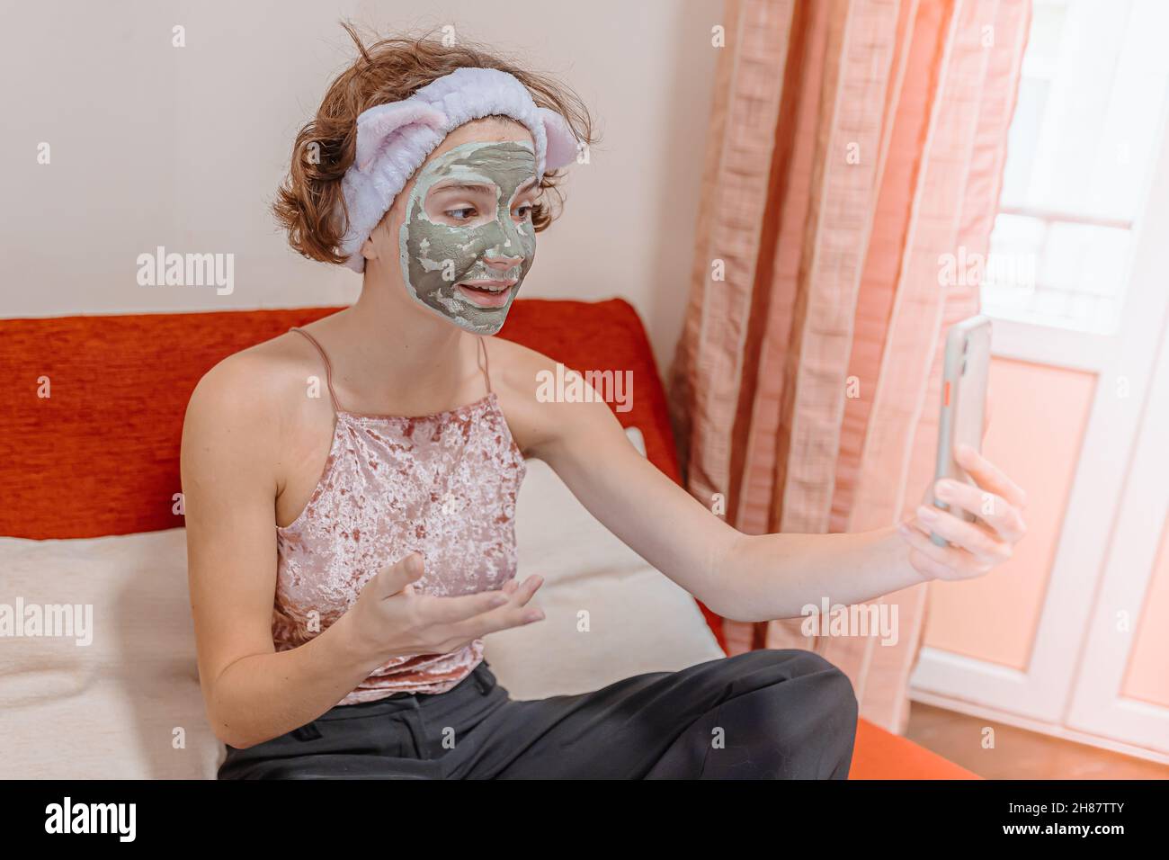 teenage girl brun curly cheveux argile masque cosmétique sur son visage prend soin de son visage à la maison et communique en ligne via le lien vidéo de sa petite amie Banque D'Images