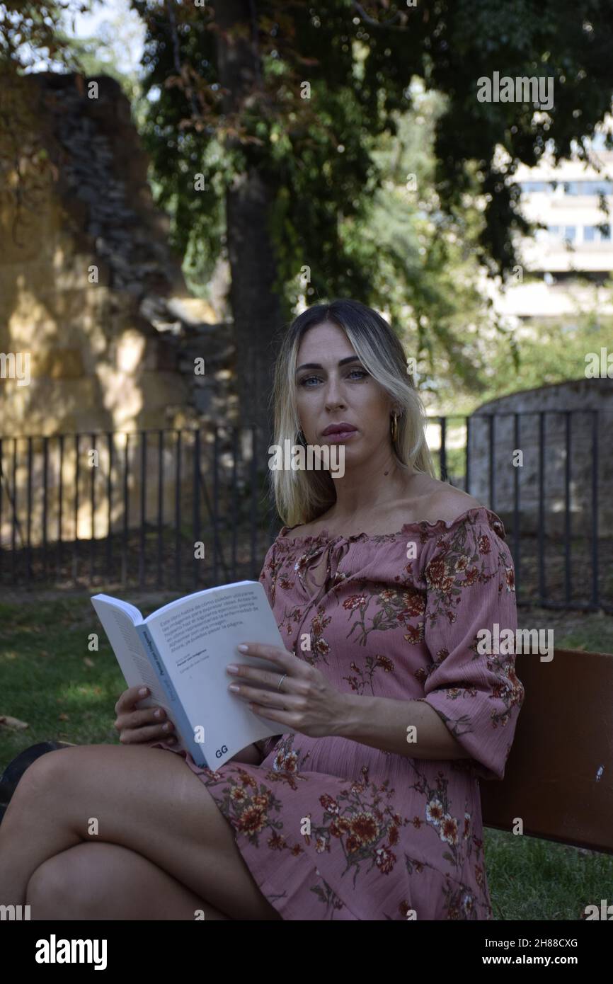 femme lisant un livre dans un parc pendant une journée ensoleillée regardant la caméra, regardant le livre, assis sur un banc, pendant un hiver, jour d'été Banque D'Images