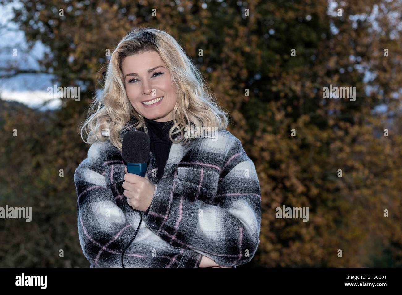 journaliste blond en manteau et microphone souriant à la caméra Banque D'Images