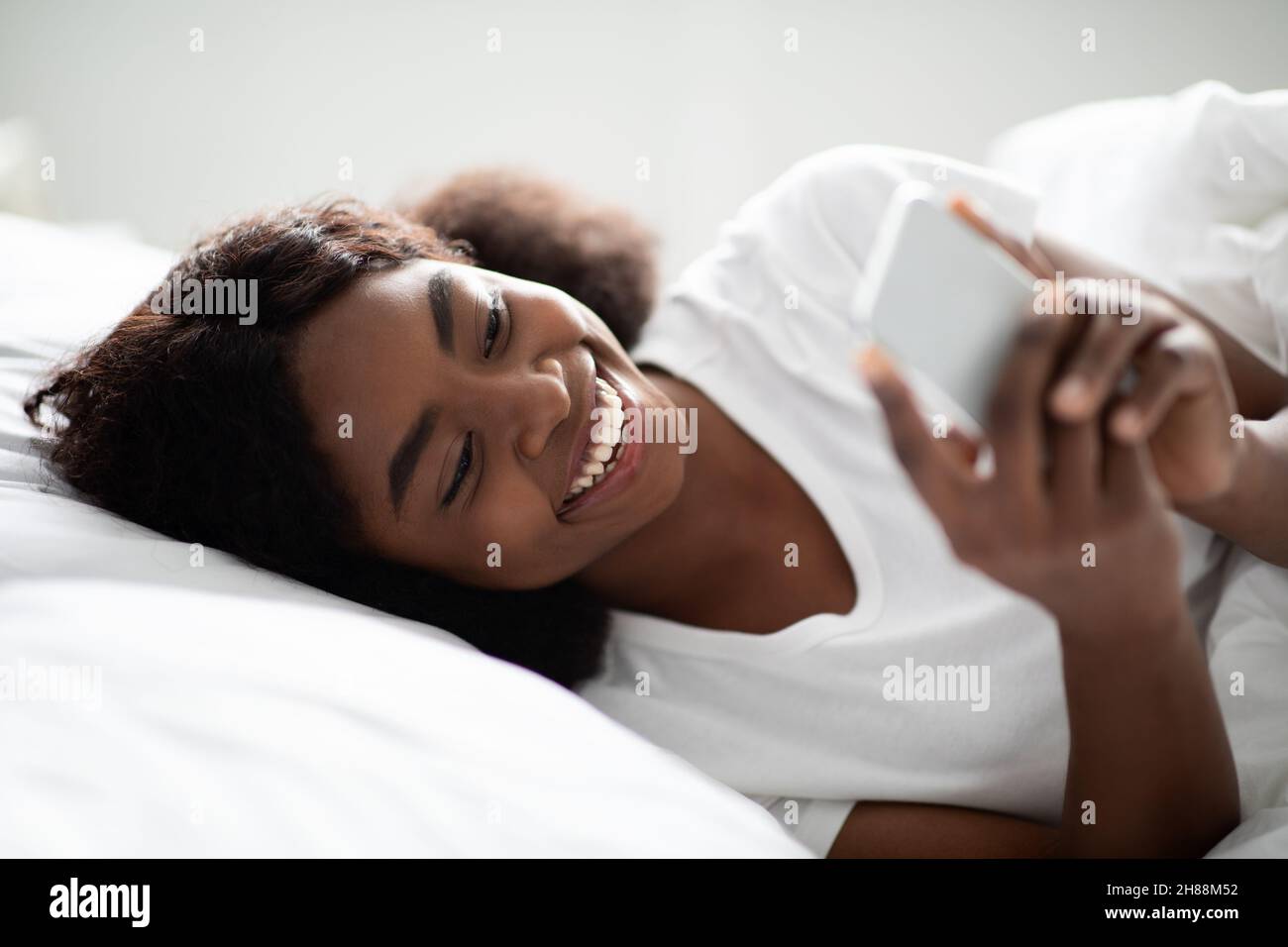 Femme afro-américaine joyeuse utilisant un téléphone portable dans le lit Banque D'Images