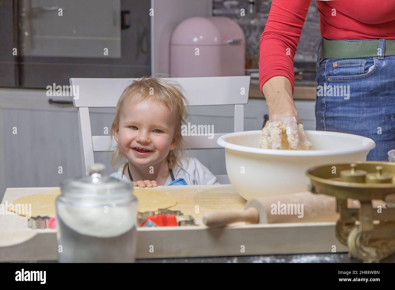 Petit chef.Le mignon petit garçon rit à l'appareil photo tandis que sa mère pétrit la pâte dans la cuisine. Banque D'Images