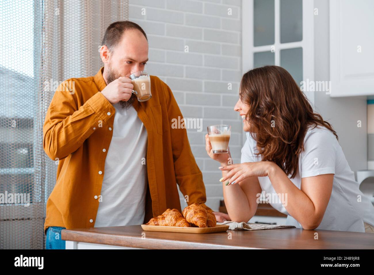 Couple de jeunes souriants homme et femme prenant le petit déjeuner dans la cuisine à la maison.Couple marié qui boit du café et qui mange des croissants dans leur Banque D'Images