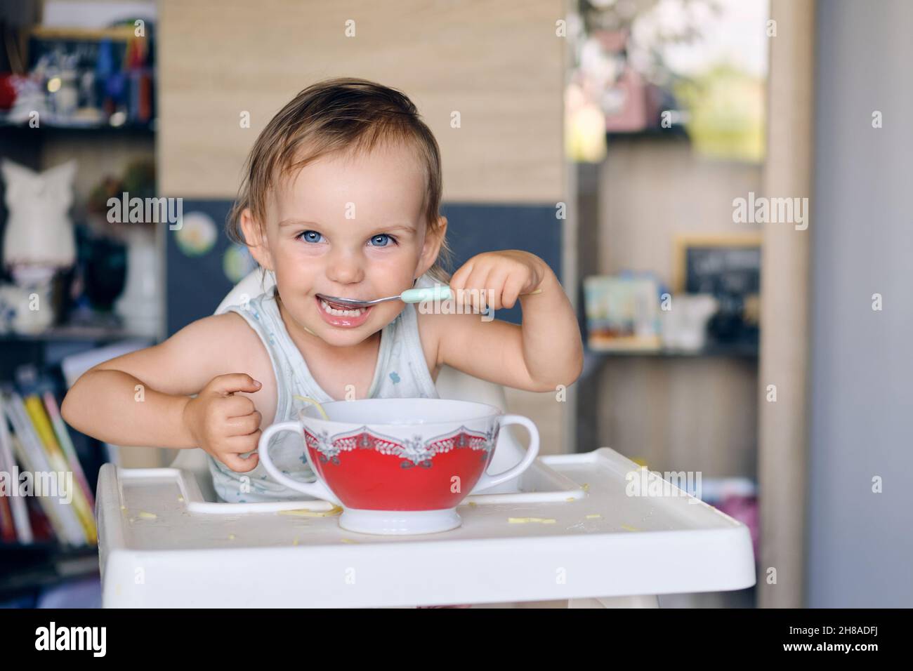 Portrait d'une jolie petite fille blonde aux yeux bleus qui mange de la soupe dans un bol en céramique et regarde l'appareil photo Banque D'Images