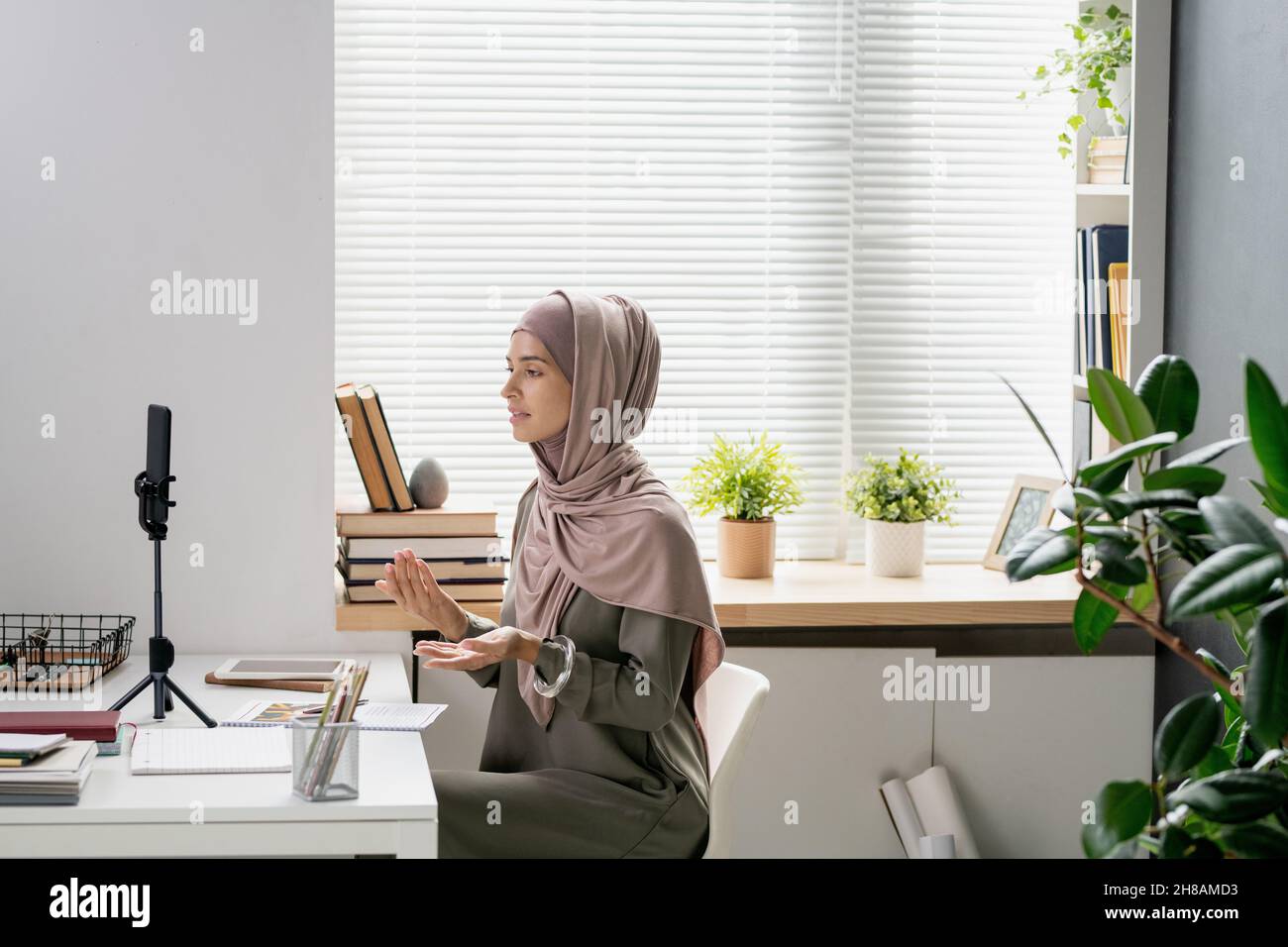 Jeune professeur confiant en hijab regardant dans l'appareil photo de smartphone tout en expliquant quelque chose aux étudiants pendant la diffusion en direct Banque D'Images