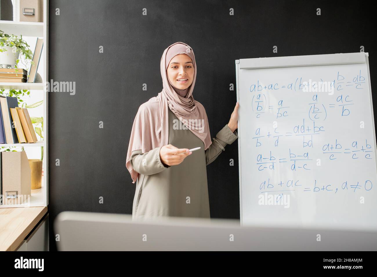 Joyeux jeune professeur de hijab debout par tableau blanc avec formule algébrique et équations tout en les expliquant devant l'ordinateur Banque D'Images