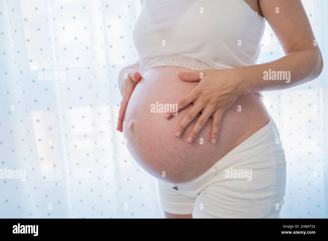 Crop anonyme femme enceinte frottant ventre près de la fenêtre Banque D'Images