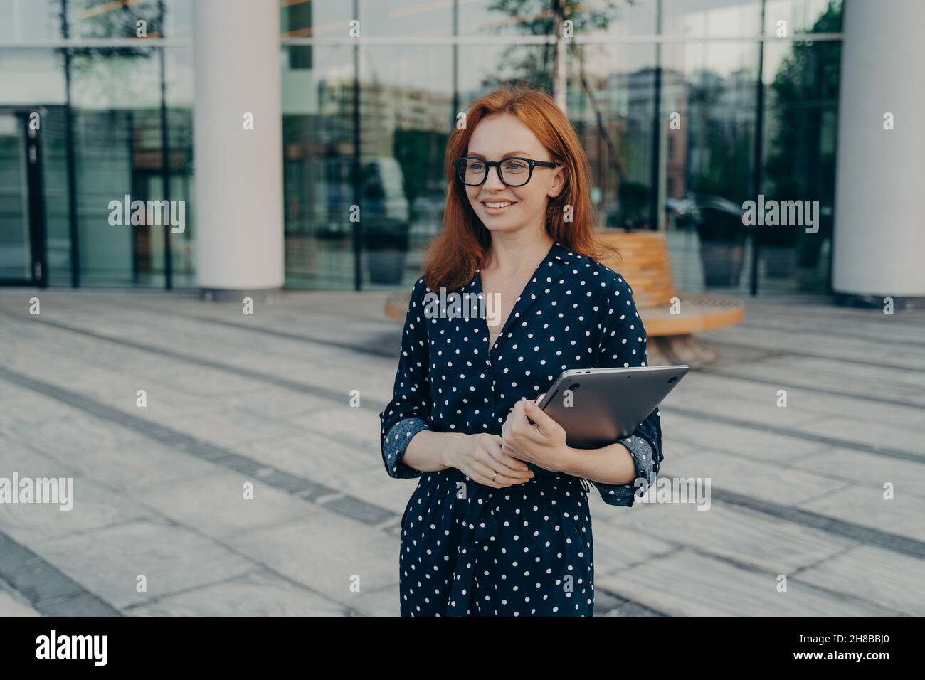 Une femme européenne avec cheveux rouges porte des lunettes et une robe à pois tient un ordinateur portable moderne Banque D'Images