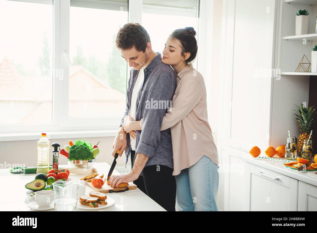 Une jeune amie embrasse son petit ami pendant qu'il coupe du pain dans la cuisine.Des aliments sains végétariens.Cuisiner ensemble.Un mode de vie sain.Famille Banque D'Images