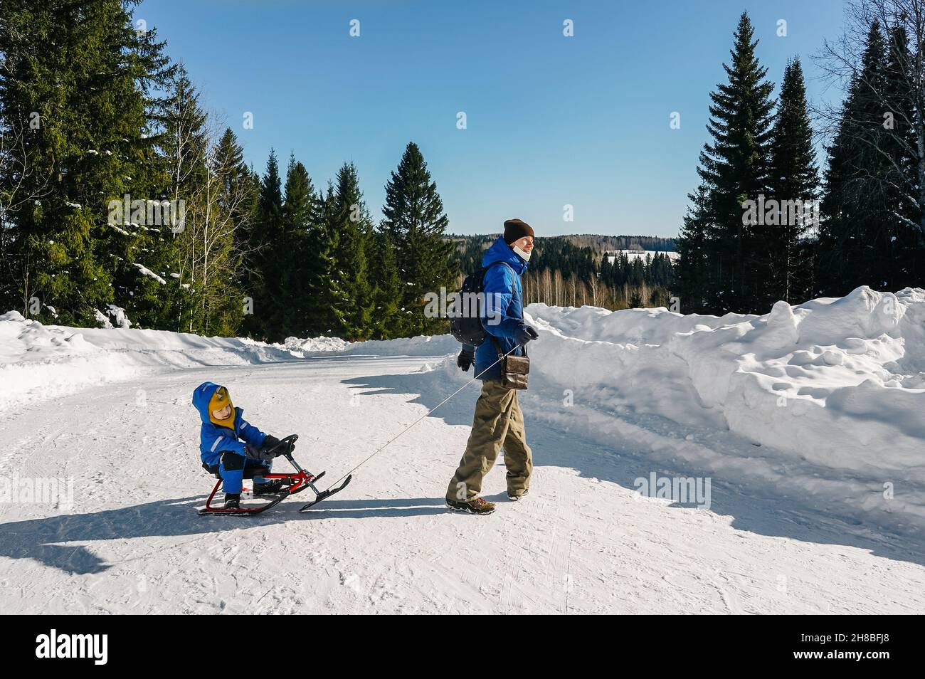 Un homme court et roule un petit garçon sur une motoneige à travers la neige sur la route forestière.Vue avant. Banque D'Images