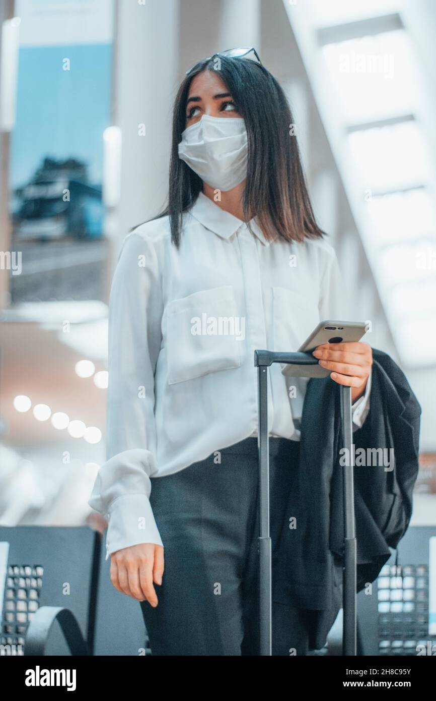 Jeune femme aux cheveux bruns avec masque facial attendant à l'aéroport avec sa valise à bagages Banque D'Images