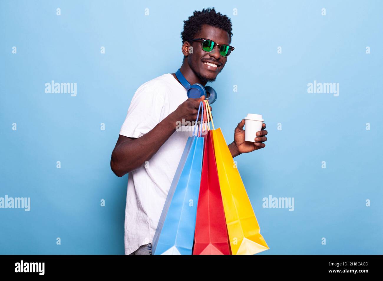 Portrait de l'homme moderne tenant des sacs de shopping et une boisson de café à la main.Personne insouciante portant des lunettes de soleil et souriant tout en portant des sacs à main après avoir acheté des vêtements en vente.Divertissement au détail Banque D'Images