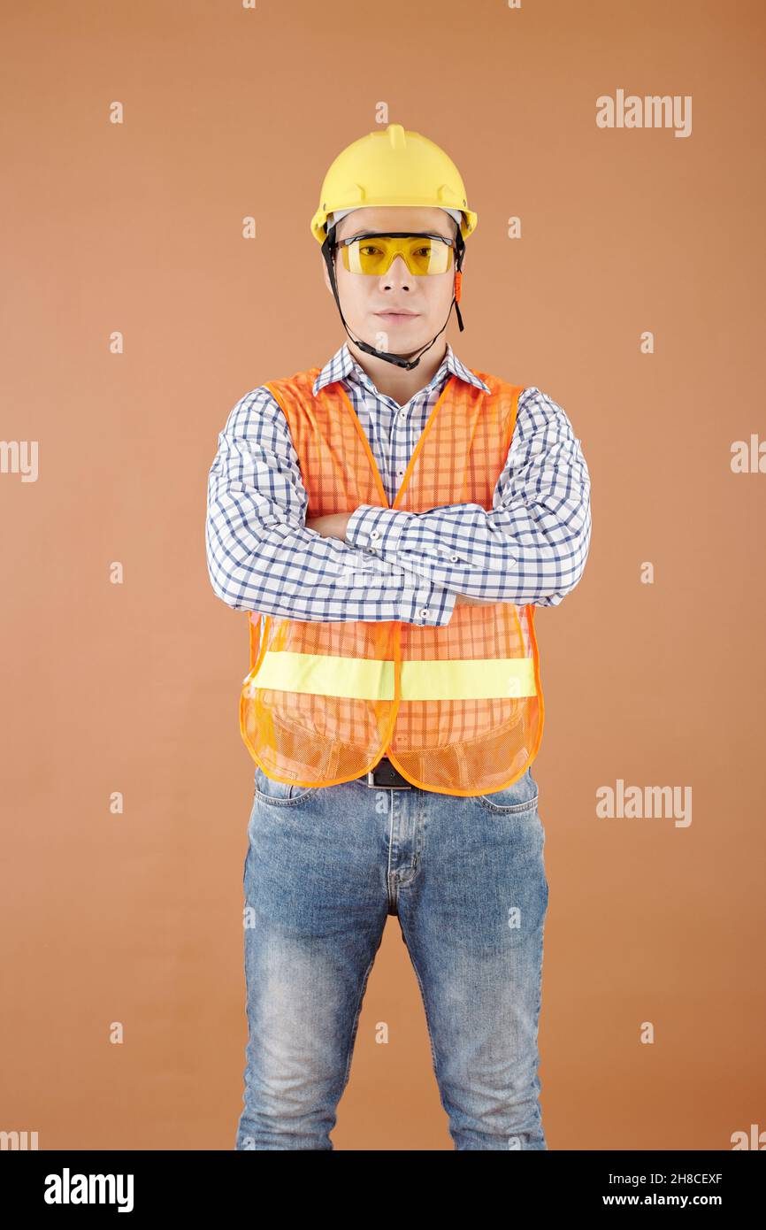 Portrait d'un entrepreneur confiant dans un gilet brillant, des lunettes et un casque de sécurité croisant les bras et regardant la caméra Banque D'Images