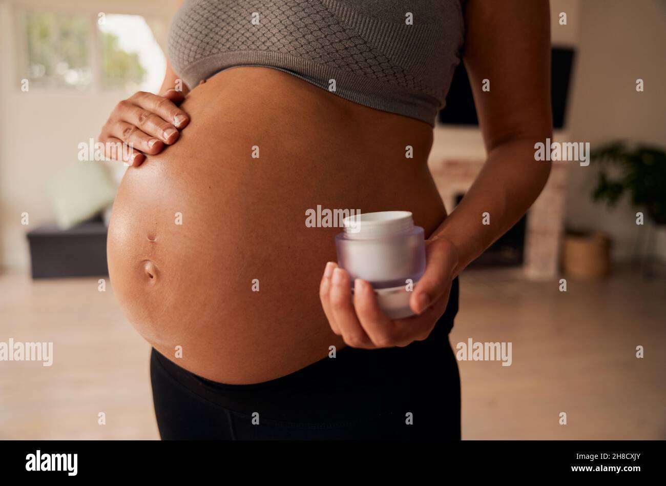 Femme caucasienne debout dans la salle de séjour avec un hydratant frottant sur le ventre de la femme enceinte Banque D'Images