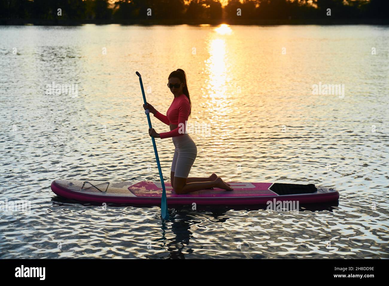 Une jeune femme en bonne santé et en forme dans des vêtements de sport et  des lunettes de soleil surfant sur le paddle board pendant le coucher du  soleil d'été au-dessus du
