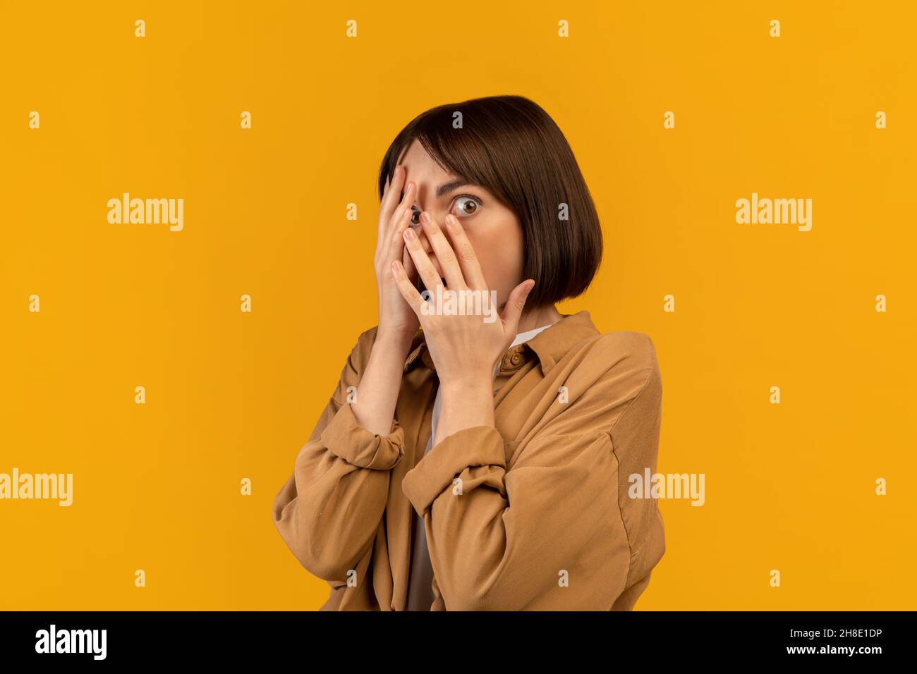 Femme effrayée couvrant le visage avec les mains et peeking avec un oeil, se sentant peur, debout sur fond jaune Banque D'Images