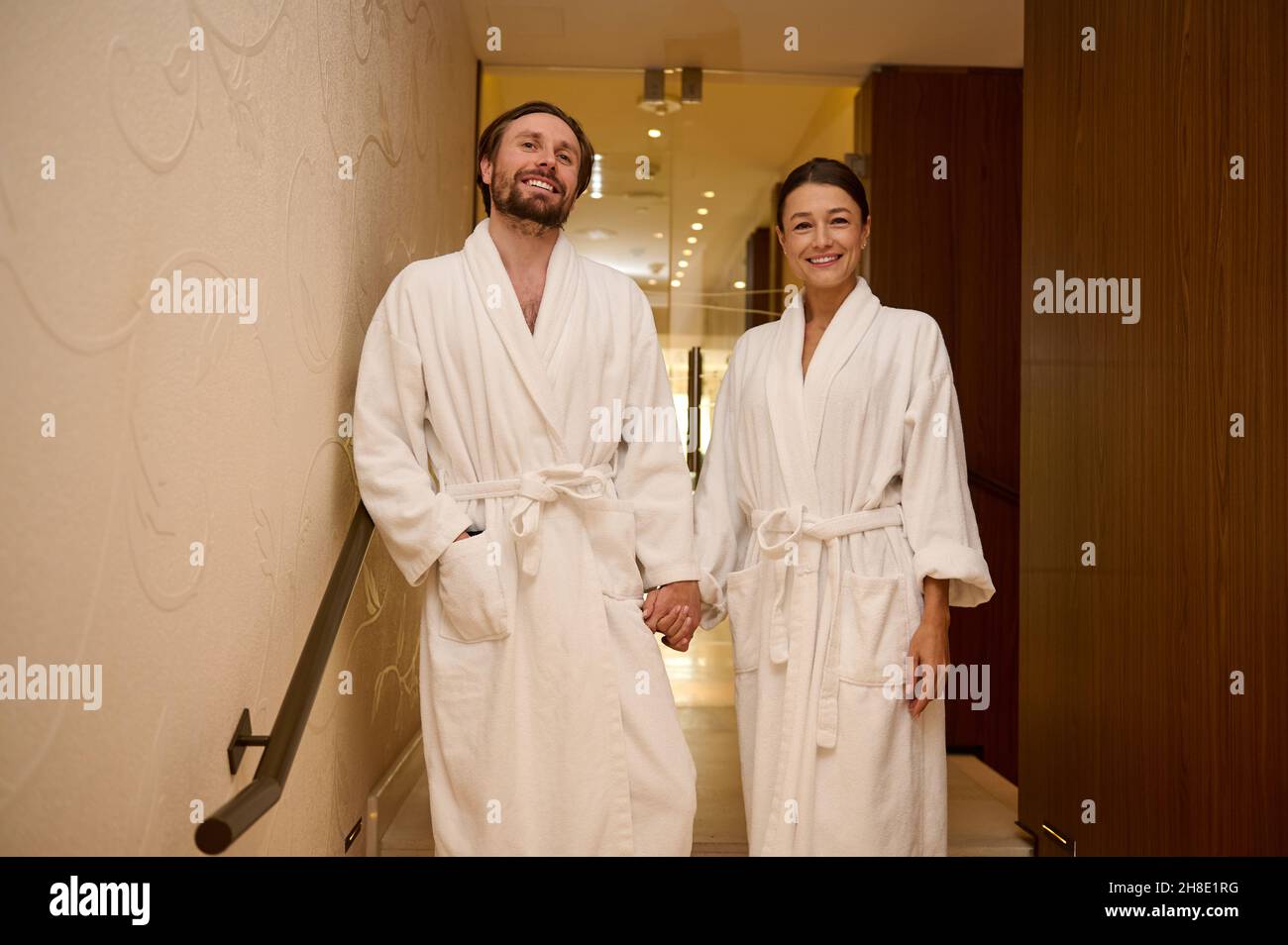 Beau couple affectueux, portant des peignoirs blancs, profitant de leur lune de miel ensemble dans un luxueux spa, en tenant les mains pendant qu'ils descendent le Banque D'Images