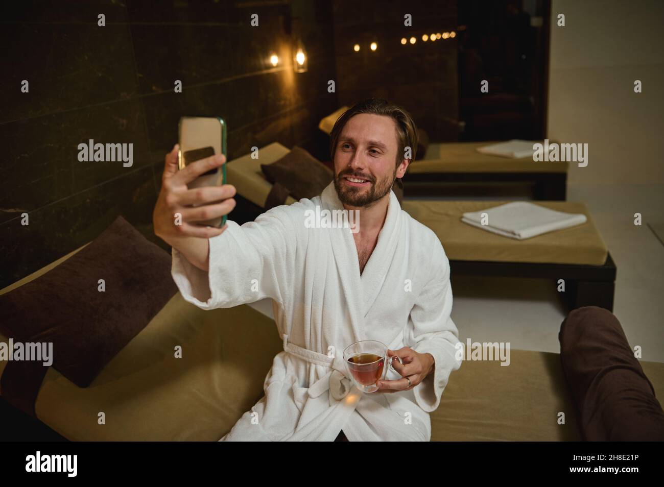 Beau homme attrayant en peignoir, assis près de la piscine dans le centre de bien-être spa, tenant une tasse de thé et sourires heureux de faire autoportrait ou parler Banque D'Images