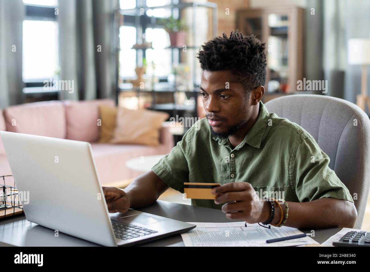 Un jeune homme afro-américain sérieux, assis à la table avec des papiers et utilisant un ordinateur portable et une carte de crédit tout en payant des impôts en ligne Banque D'Images