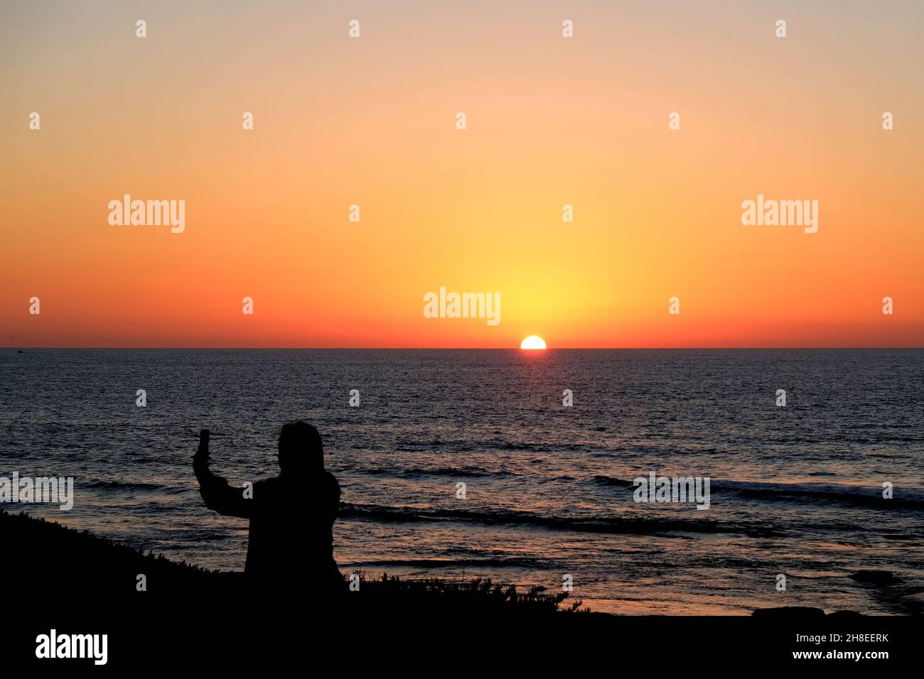 Une jeune femme prenant un selfie capturant le moment où le soleil se couche dans l'océan Pacifique à la Jolla, Californie, États-Unis ; coucher de soleil à San Diego. Banque D'Images