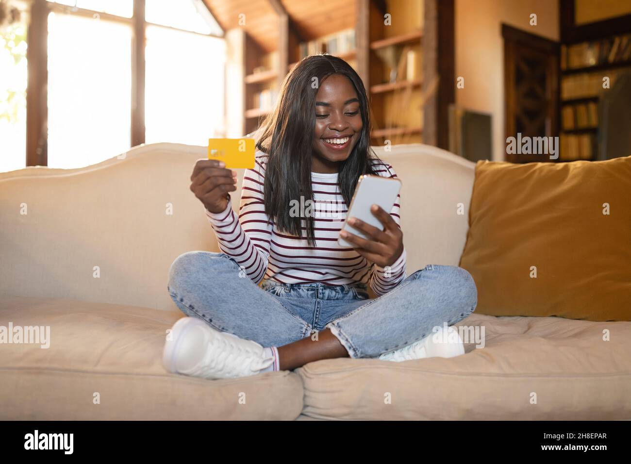 Jeune femme noire avec carte de crédit et smartphone assis à pieds croisés sur le canapé, magasiner en ligne de la maison Banque D'Images