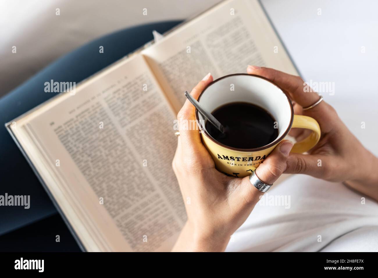 Détail des mains d'une fille couchée au lit avec une tasse de café dans ses mains et un livre Banque D'Images