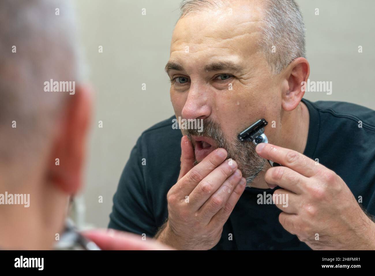 Homme mûr aux cheveux gris recoiffant lui-même sa barbe à la maison à l'aide d'un rasoir Banque D'Images