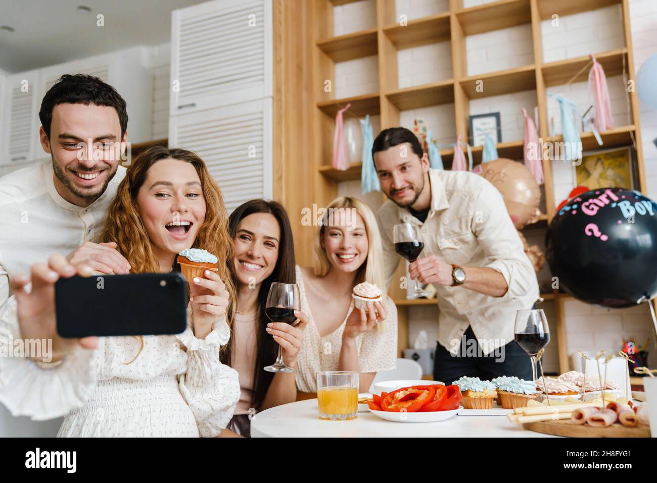 Bonne jeune femme enceinte célébrant la douche de bébé avec ses amis à la maison, prenant selfie ensemble, debout à la table Banque D'Images