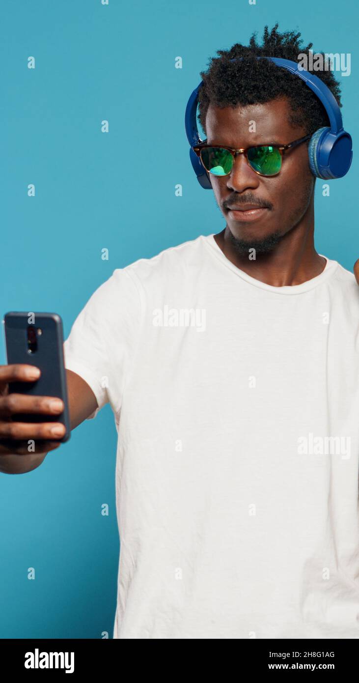 Homme espiègle prenant des photos avec des sacs d'achats sur un smartphone tout en portant des lunettes de soleil et des écouteurs. Adulte moderne prenant des selfies avec le téléphone après l'achat de vêtements dans la vente au détail. Banque D'Images