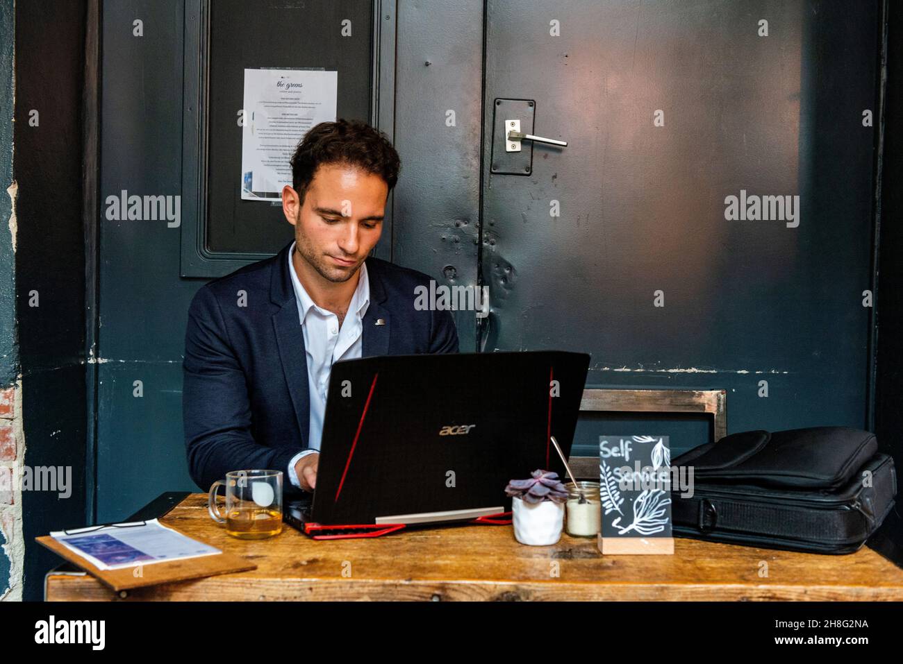 Berlin, Allemagne. Jeune adulte et professionnel gay travaillant avec son ordinateur portable et son smartphone à l'intérieur d'un café pour aider ses clients avec leur marketing en ligne et seo. Banque D'Images