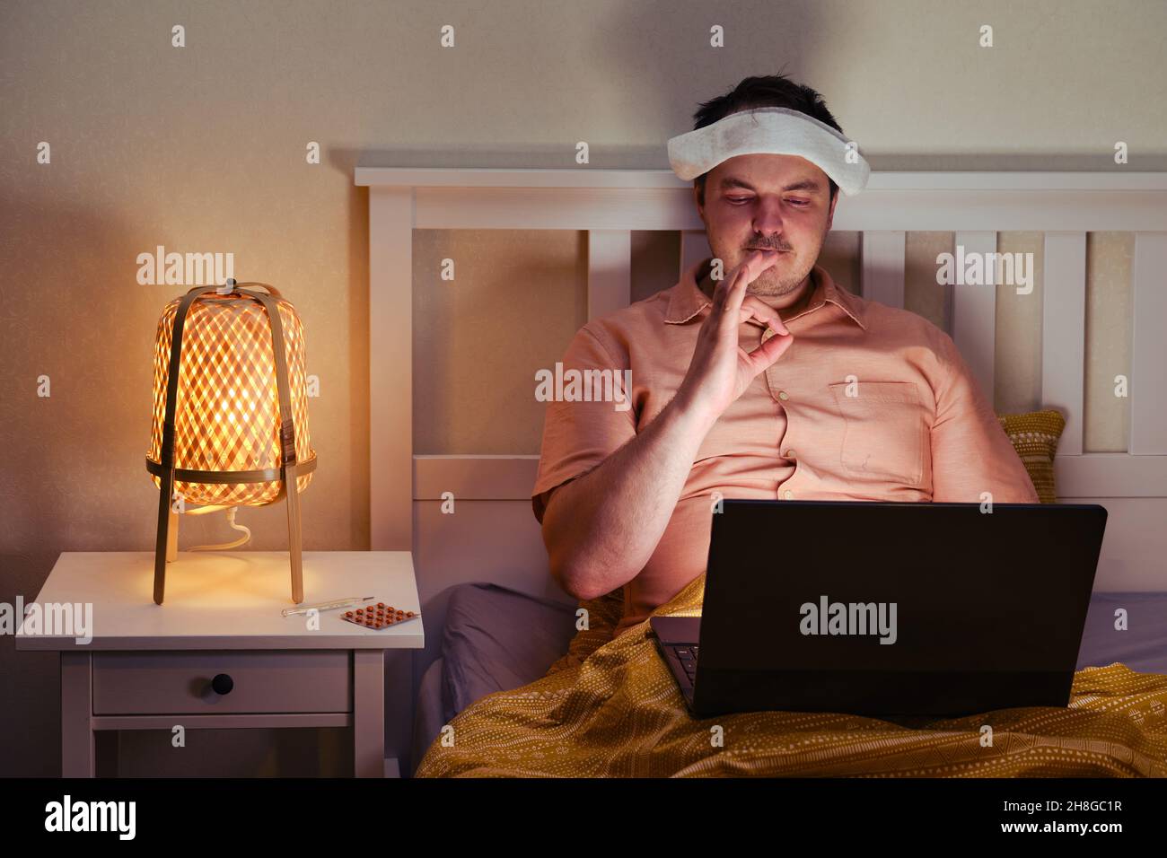 Un homme malade adulte montre un geste acceptable à l'écran avec un ordinateur portable sur le lit à la lumière d'une lampe de nuit Banque D'Images